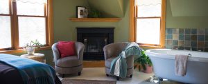 large guest room with green walls, corner fireplace, two gray upholstered chairs, blue bathroom and large clawfoot tub
