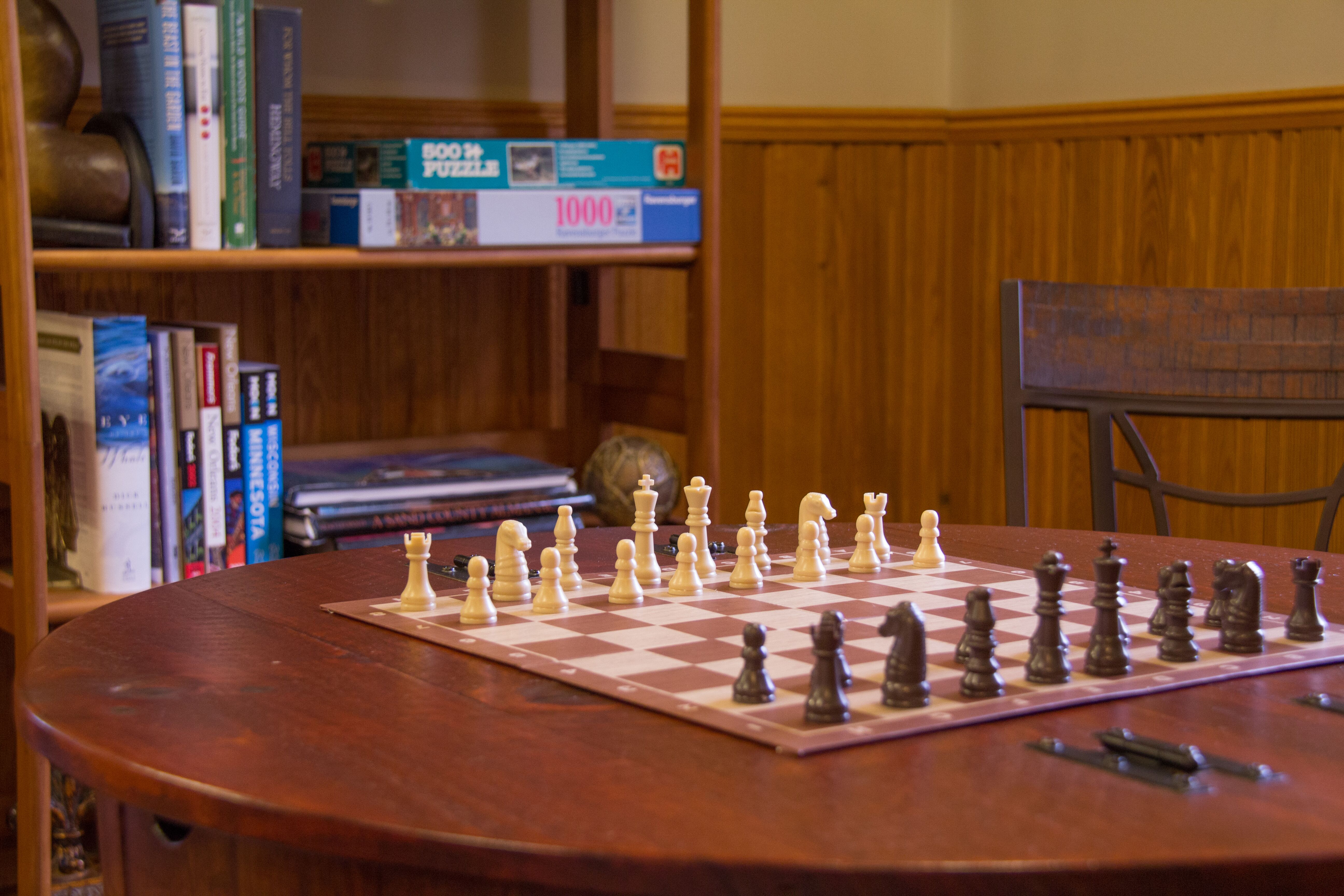 chess set on wood table with chair and shelves with books and puzzles in background