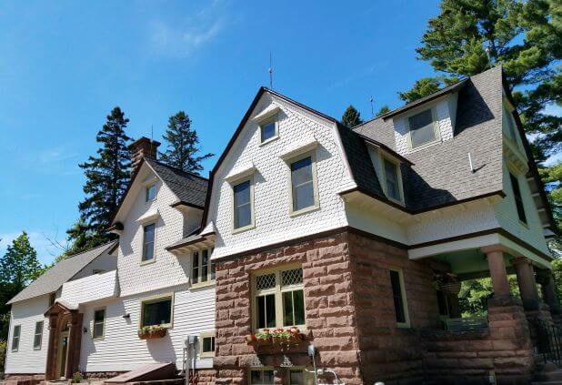 brownstone and tan wood siding house with blue sky and tall pine trees