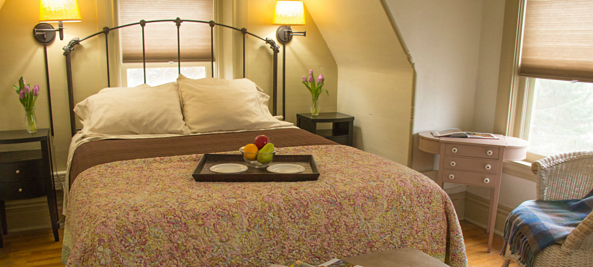 Beige guest room with wood floors, sloped ceiling, metal bed flanked by nightstands and reading lights and two windows