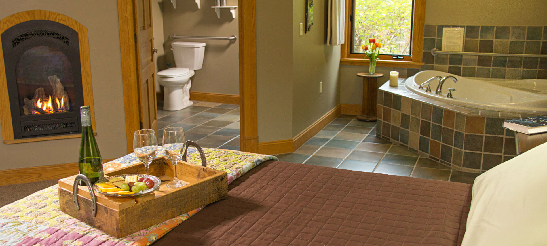 Tan guest room with fireplace, bed topped with tray of wine, cheese, crackers and grapes, and view of corner soaking tub