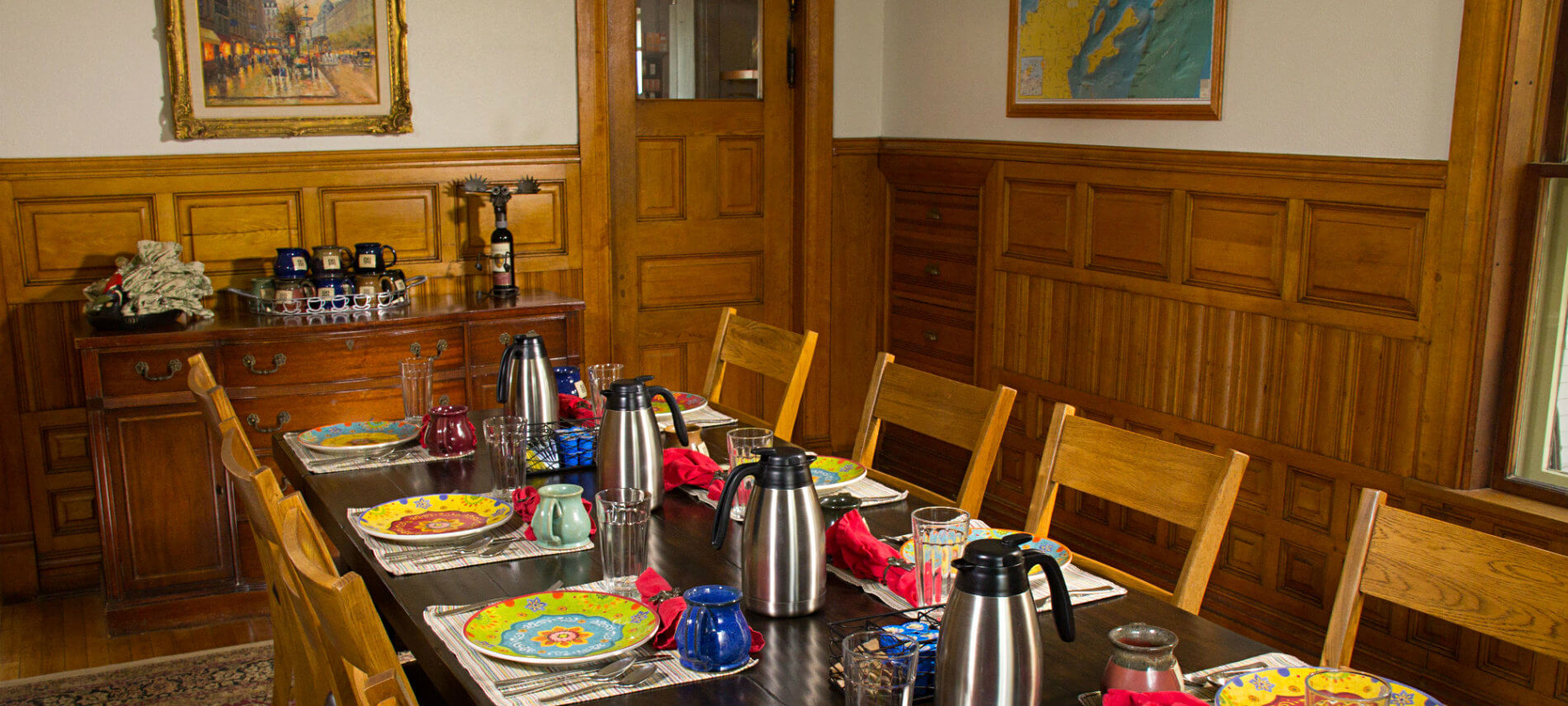 Dining room with wood floors and wainscotting, and long wood table set for breakfast with colorful plates