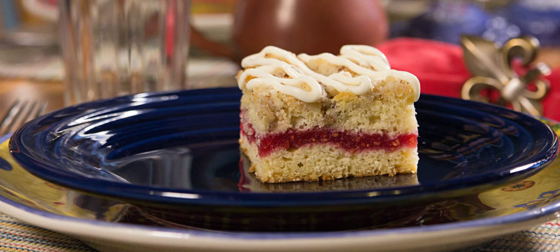 Blue ceramic plate on a colorful ceramic charger topped with a piece of raspberry coffee cake drizzled with icing
