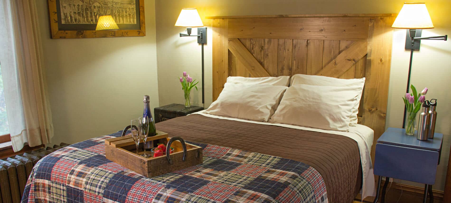 Beige guest room, bed with plaid quilt and rustic wood headboard, tray of strawberries and champagne, nightstands and reading lights