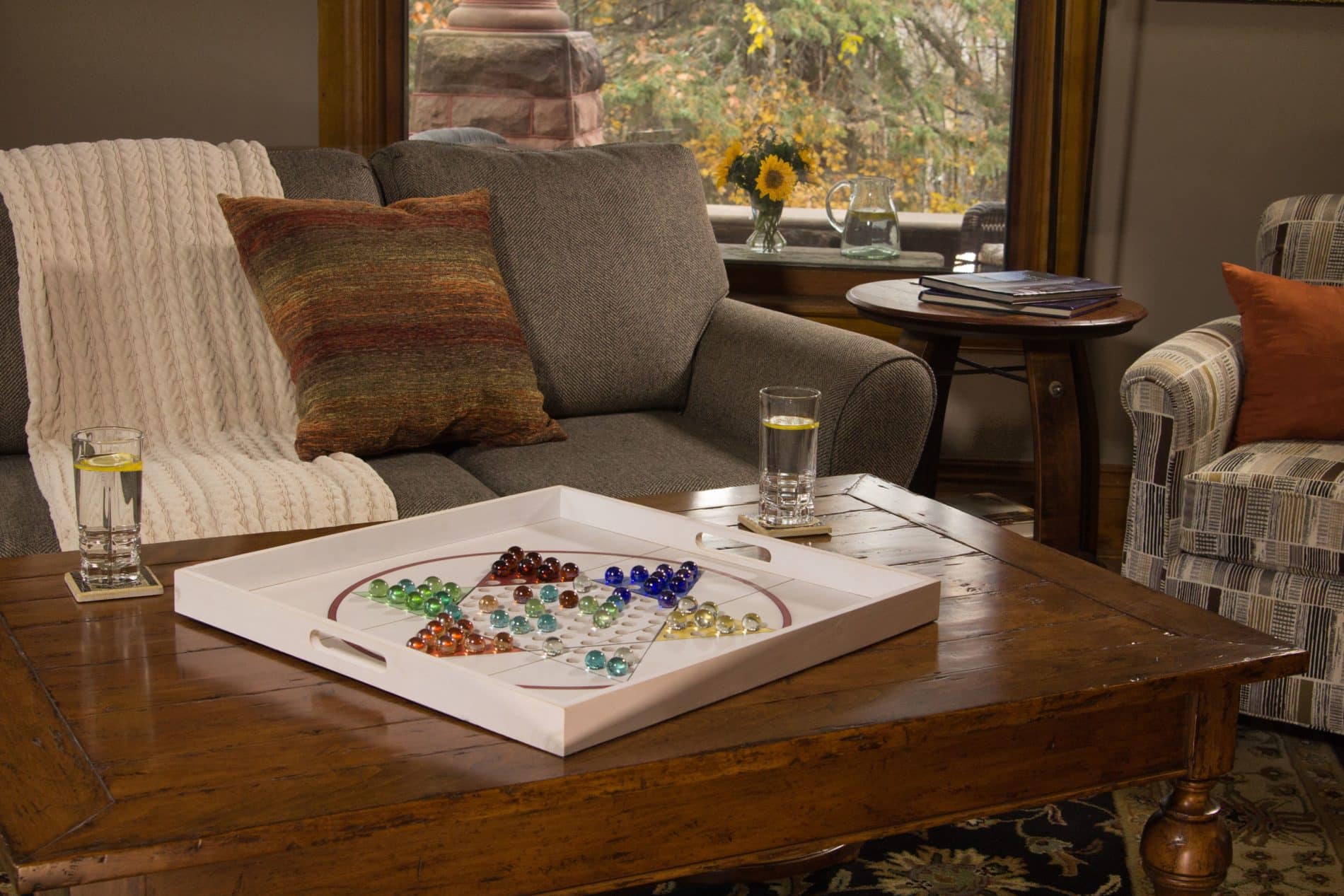 gray couch with multi color throw pillow and wood coffee table with large white chinese checkers board and two glass of water with lemon on top