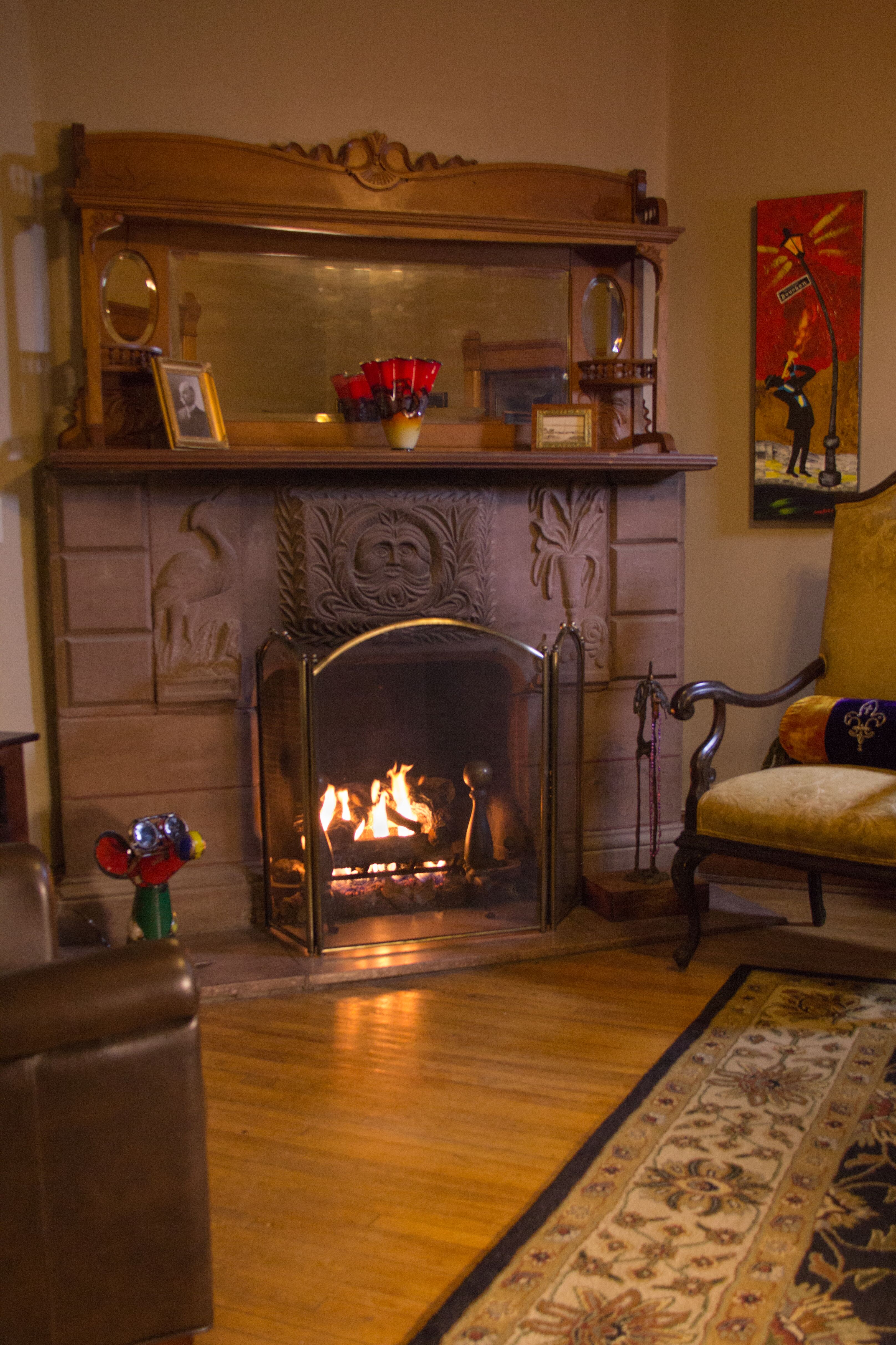 brownstone carved fireplace with roaring fire with mantle and mirror, a colorful metal mouse with glasses and a gold antique chair off to the side