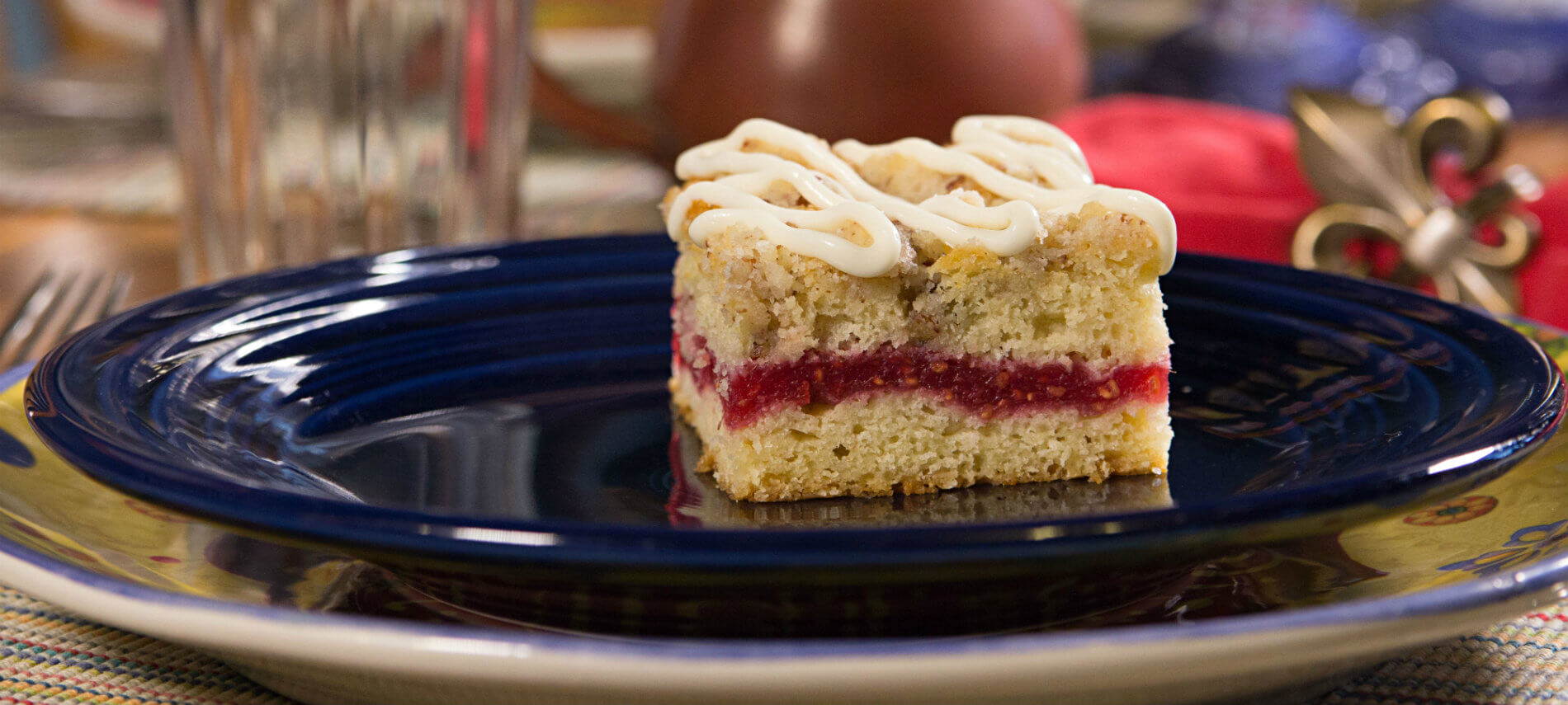 Blue ceramic plate on a colorful ceramic charger topped with a piece of raspberry coffee cake drizzled with icing