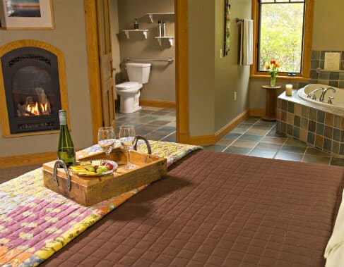 Tan guest room with fireplace, bed topped with tray of wine, cheese, crackers and grapes, and view of corner spa tub