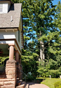 Exterior side view of inn with covered porch, stone pillars, surrounded by green grass, tall green trees and blue skies